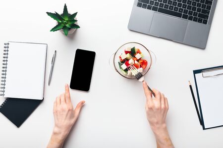 Healthy eating concept. Woman eating fresh salad and looking at cellphone with blank screen, work at home conceptの写真素材