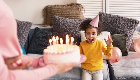 Little african american girl in cap rejoices to cake. Birthday at home during quarantine