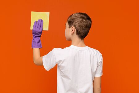 Home cleaning. Teen boy washing wall with sponge, orange studio background, back view