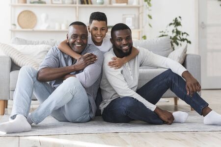 Three Generations Of Men. Smiling Black Boy, Father And Grandpa Posing To Camera While Sitting On Floor At Homeの写真素材