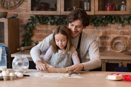Cute little girl and her father in aprons rolling out the dough together on kitchen table, making cookies, free spaceの写真素材