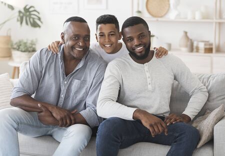 Male Generation. Smiling Black Boy Embracing His Dad And Grandfather Posing To Camera At Home, Sitting Together On Couch In Living Room.の写真素材