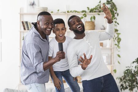 Cheerful Black Father, Son And Grandfather Having Fun At Home, Singing Together In Living Room, Using Remote Controller As Microphone, Free Spaceの写真素材