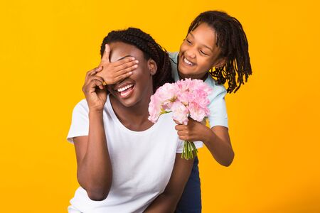 Mothers Day Concept. Black child keeping her moms eyes covered and holding flowers, yellow studio wallの写真素材