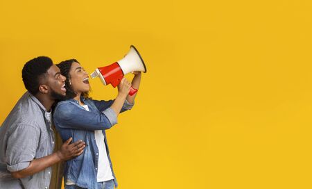 Making announcement. Positive black couple shouting in megaphone at copy space over yellow background, panoramaの写真素材