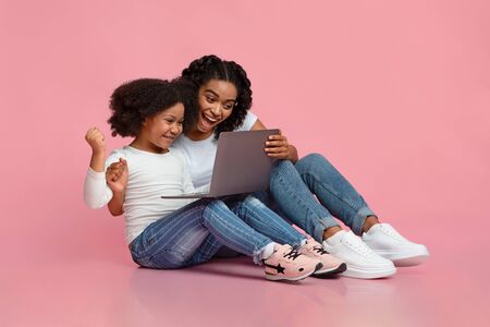 Overjoyed Black Mom And Little Daughter Celebrating Success With Laptop, Sitting Together On Floor...