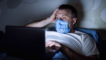 Coronavirus Panic. Scared Mature Guy In Mask Reading News Online On Laptop Lying In Bed Indoors...