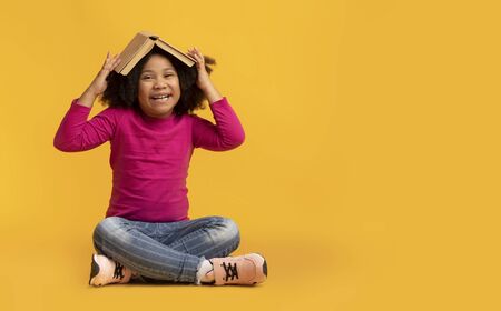 Education Concept. Portrait Of Playful Little Afro Girl With Opened Book On Head Sitting On Yellow Background In Studio And Laughing, Copy Spaceの写真素材