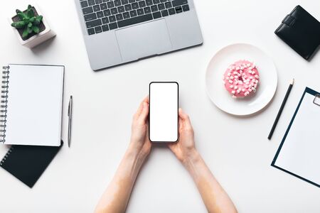 Feminine desktop. Female hands holding cellphone with blank screen and morning donut on plate, copy spaceの写真素材