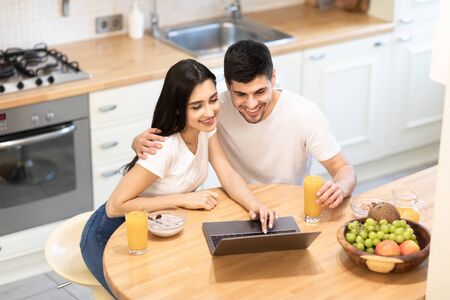 Romantic young couple having breakfast and using laptop at table in modern kitchen, drinking orange juice, high angle viewの写真素材