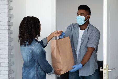 Food delivery during coronavirus. Black courier guy wearing medical mask delivering grocery order to young womans homeの写真素材