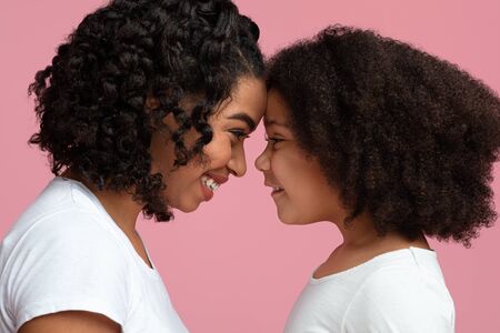 Cute little black girl touching foreheads with her loving mother, looking in her eyes, standing over pink background, side viewの写真素材