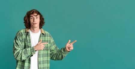 Surprised teenage guy pointing to empty space on turquoise studio background