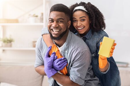 Loving Black Couple Cleaning Flat Together, Embracing And Posing To Camera, Holding Sponge And Wearing Rubber Gloves, Copy Spaceの写真素材