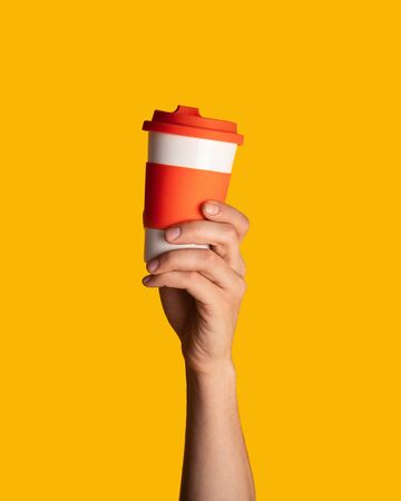 Young guy holding reusable eco mug with coffee on orange background, close up