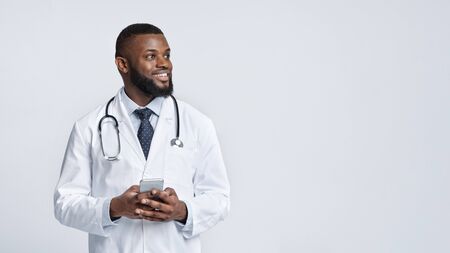 Call your doctor during quarantine. Happy african doctor with mobile phone looking at empty space over white studio background, panoramaの写真素材