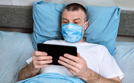 Coronavirus Quarantine. Middle-Aged Man In Mask Using Digital Tablet Watching Movie Lying In Bed At...