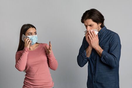 Coronavirus Symptoms. Concerned Young Woman In Medical Face Mask Talking On Cellphone Next To Her Sick Boyfriend, Calling To Doctor Or Emergencyの写真素材