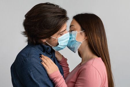 Love During Epidemy. Millennial Couple Kissing Each Other While Wearing Medical Face Masks, Standing...