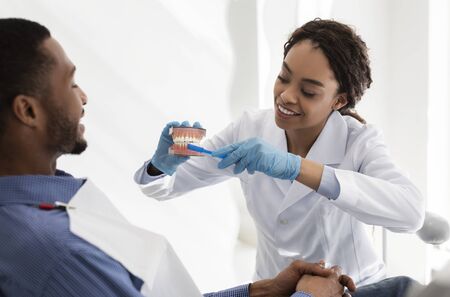 Positive african woman dentist doctor explaining patient how to brush teeth, using toothbrush and...