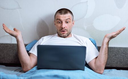 Computer Problem. Displeased Man Using Laptop Shrugging Shoulders Having Issue With Internet Connection Sitting In Bed At Home.の写真素材