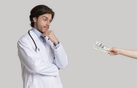 Doubtful young doctor looking at money bribe offered by female patient, thoughtfully touching chin while standing on light background in studioの写真素材