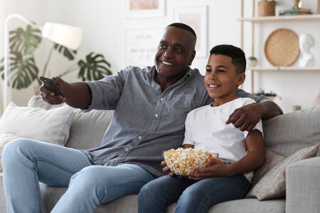 Home Entertainment. Happy African American Grandfather Watching TV With Grandson In Living Room And Eating Popcorn Together, Relaxing On Couchの写真素材