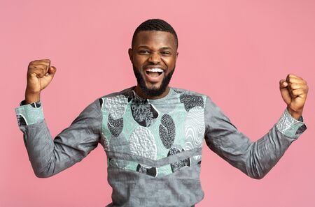 Excited african man in traditional blue shirt screaming and clenching fists over pink background