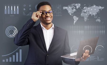 Online business concept. Happy Black Businessman In Digital Glasses Holding Laptop, grey background with business bars and schemesの写真素材