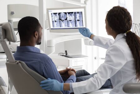 Female dentist african woman pointing at screen with xray picture, showing patient his teethの写真素材