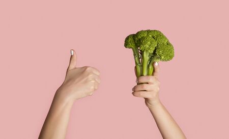 Healthy eating and dieting concept. Young girl holding broccoli and showing thumb up gesture on pink background, close up. Panoramaの写真素材