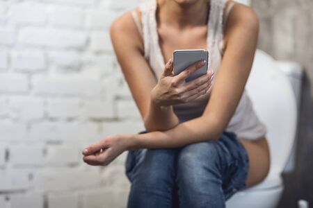 Unrecognizable Girl Using Cellphone Sitting On Toilet Having Constipation Problem In Restroom Indoors. Defecation Concept. Cropped, Empty Spaceの写真素材