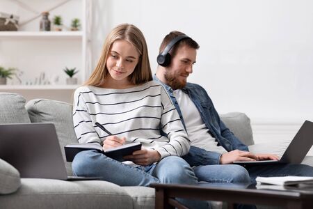 Millennial lifestyle. Young couple using laptops at home, girl studying and guy playing video gameの写真素材