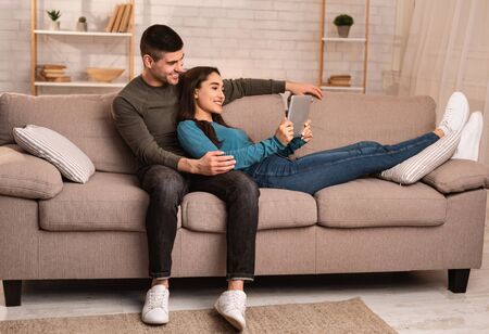 Stay At Home Concept. Young lovely couple spending time together at home indoors, using tablet, lying on sofaの写真素材