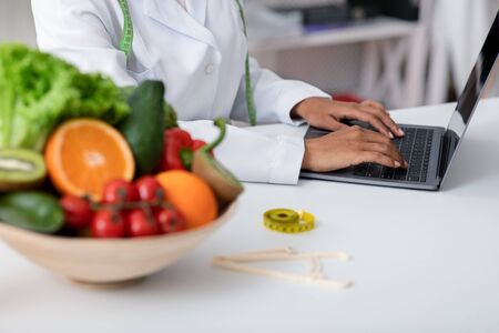 Cropped of fresh fruits and vegetables bowl next to working online african doctor, dietician using laptopの写真素材