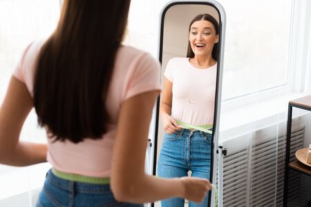 Weight Loss. Happy Girl Measuring Waist With Tape Standing In Front Of Mirror Slimming At Home. Copy...