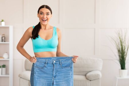 Successful Weight Loss. Thin Girl Comparing Old Oversize Jeans After Dieting Standing Indoors. Copy Spaceの写真素材