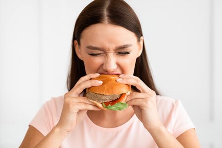 Cheat Meal. Hungry Girl Biting Burger Enjoying Eating Junk Food Having Diet Break Standing At Home.