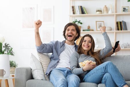 Emotional man and woman watching football game together at home, exclaiming happiness, eating...