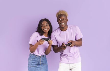 Cheerful African American couple with joysticks playing video games on lilac background