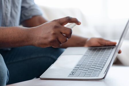 Closeup Of Black Man Using Disinfectant Spray On Laptop Keyboard Before Use At Home, Cropped Image