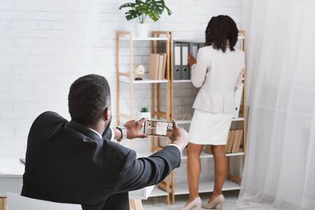 Sexual molestation in office concept. Back view of black guy taking photo of his female coworker at workplaceの写真素材