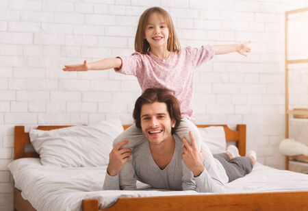 Father and daughter spending time together at home in morning. Happy little girl sitting on father shoulders with hands up, dad laying on bed, free spaceの写真素材