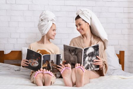 Beautiful young woman and her teen daughter reading fashion magazines and doing nails after bath in bedroomの写真素材