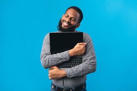 Technology Lover Concept. Happy black man hugging laptop with blank screen, isolated over blue studio wallの写真素材