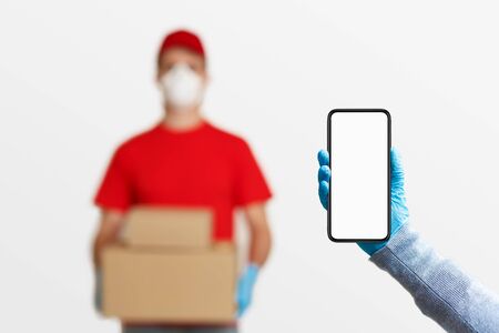 Delivery of packages on time concept, and hand with smartphone with blank screen. Blurred courier in protective mask and gloves with parcelsの写真素材
