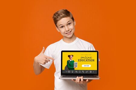 Cheerful boy pointing at laptop with online education site on screen, orange background. Creative...