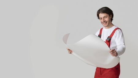 Young Repairing Worker Checking Blueprint, Holding Project Plan In Hands And Smiling, Posing Over Light Studio Background, Panorama With Free Spaceの写真素材