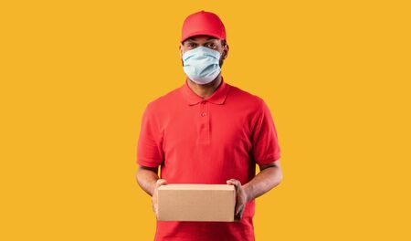Delivery Service. African Courier Guy Holding Cardboard Box Delivering Parcel Package Wearing Protective Mask Posing Over Yellow Background. Studio Shotの写真素材