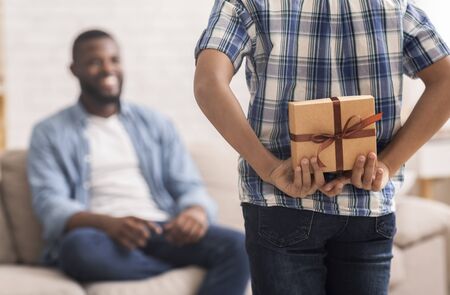 Boy greeting his black dad with Fathers Day. Little child hiding gift box behind his back, selective focusの写真素材